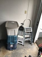 Photo showing four step stools of various sizes and three large storage totes stacked, positioned near a wall with an electrical outlet, in a garage or utility space.