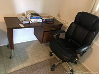 Desk and chair side view showing the laminate desk with books and puzzle on top and a black padded swivel chair beside it.