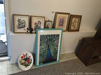 Full view of seven framed art pieces including shadow box children's art, peacock puzzle, flower plaque, and fruit print arranged near a wall.