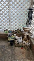 Various garden items arranged near lattice backdrop including animals, planters, pedestal, and a cardboard box