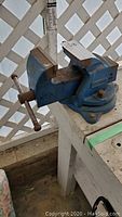 Front and side view of the blue Record No. 3 bench vice mounted on a wooden workbench with a metal handle for clamping.
