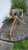 Yellow wheelbarrow with various garden tools including pitchfork, spades, rake and shovel arranged inside and on gravel surface.