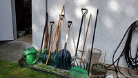 Photo of garden and hand tools including three snow shovels, watering can, leaf rake and dirt rake against an exterior wall.