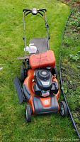 Overview of mower with fuel can on top, grass catcher attached, spare blades and fuel jug on ground