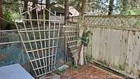 Two wooden garden trellises leaning against a wire mesh fence outdoors near a wooden fence, surrounded by stones and ground debris.