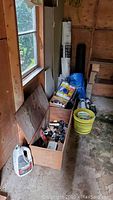 View of wooden box filled with plumbing fittings, tools, and other hardware supplies near a window and buckets on the floor.
