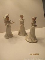 Photo showing three Lenox Victorian lady figurines each in white and gold trimmed Victorian dresses, one holding a parasol, one a croquet mallet, and one holding a bouquet.