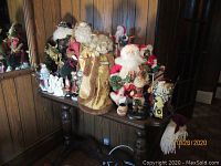 Multiple Santa Claus figurines displayed on a table against a wood-paneled wall, showcasing variety in size, clothing, and style.