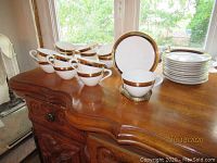 Set of cups, plates and bowls with platinum and gold bands on white porcelain, arranged on wooden surface near window.