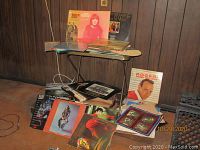 Multiple vinyl records and album covers stacked and displayed on and around a small folding table in a wood-paneled room. Visible album covers include Roger Williams, Andy Williams, Johnny Mann Singers, and Rogers & West.