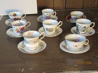 Wide view of all 8 collectible cups and saucers arranged on a table, showing floral and gold detail designs.