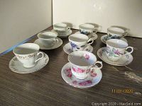 Eight assorted collectible cups and saucers arranged on a wooden surface with various floral and animal designs.