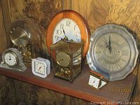 Wide view of shelf displaying seven clocks including anniversary clock in glass dome, wooden framed wall clock, metal wall clock with roman numerals, marble base clock, white plastic travel clock, small folding travel clock, and brass carriage clock.