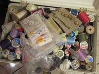 Overview of plastic container filled with assorted spools of thread, packets of white buttons and beads, small gold-tone buttons on card.