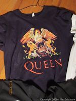 Black Queen t-shirt with colorful emblem of eagle, lions, and crown on front hung on wooden floor