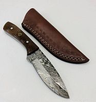 Full view of the hunting knife and accompanying leather sheath lying flat showing the Damascus steel blade pattern and the wooden handle with custom medallion rivet.