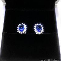 Front view of earrings showing blue tanzanite center stones with clear stone halos, displayed in a black velvet jewelry box.