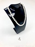 Pearl necklace and matching pearl earrings displayed on black bust stand and flat on white background.
