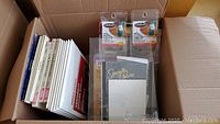Top-down view inside an open box showing a collection of photo cards and various paper packages upright, including plastic blister packs with Eldon branded products on top.