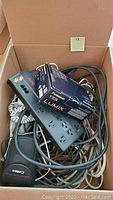 View of items including Canon camera lens, Panasonic Lumix TZ5 camera box, APC Surge Arrest power cord, cords, and other accessories in a cardboard box