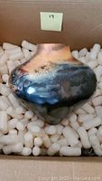 Side view of handcrafted ceramic vase showing glazed finish with tan, black, and blue tones inside a box with packing peanuts.
