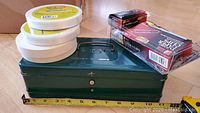 Photo showing a dark green vintage metal box with handle, flap closed, with two rolls of white masking tape and filament tape on top, along with packages of jumbo paper clips and binder clips.