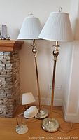 Full view of two tall brass and metal floor lamps with white pleated shades next to a stone fireplace and one smaller brass lamp on the floor, all with visible cords and plugs.
