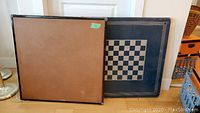 Two folding tables leaning against a wall, one plain wooden, one metal with a printed chess board.