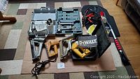 Lot includes various tools and car accessories placed on carpet and cardboard: DeWalt jig saw in metal case, staple guns, power tools, car jump cables, car mat, snow brush and sunshade visible