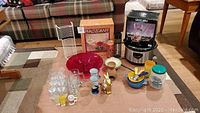 Photo showing full lot items on floor including Rival crockpot, plate rack, Pfaltzgraff glassware set, Cameleon salad set, plastic bowls, glasses, rabbit figure, and other kitchen items.