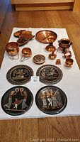 Overview of all items on white cloth, showing copper elephant clock, bell, pitch and cauldron, bracelet, trinket case, and four decorative plates with figures.