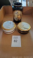 Photo of two round trinket boxes, one white and one black, and a tall dark vase with gold floral pattern, all placed in a cardboard box.
