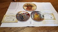 All three collectible plates displayed with certificates on a white cloth background on wooden floor.