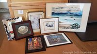 Seven framed art pieces arranged grouped together, showing variety of sizes and subjects including seascape and urban scenes.