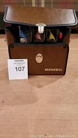 Four slot cars arranged vertically inside open brown Bushnell carrying case with silver latch on top, displayed on a tabletop.