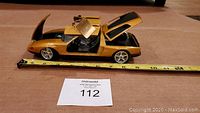 Side view of gold metallic Mercedes Benz C111 diecast model car with gull-wing doors open, showing seating interior and wheels, on measuring tape showing length about 9.5 inches.