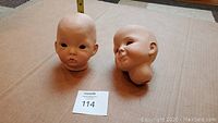 Two porcelain doll baby head sculptures on a brown cardboard surface, one looking ahead and the other head tilted with a smile.