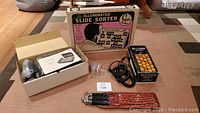 Wide shot of all items on floor showing slide sorter box, calculator, Philips tape, tripod pouch, and tape recorder.
