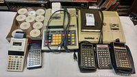 Photo showing large vintage adding machine with cord, box of paper rolls, small white adding machine, and three handheld calculators