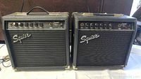 Front view of two Squier Champ 15G 15W guitar amplifiers side by side showing control panels, handles, and speaker grilles.
