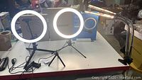 Two ring lights lit and mounted on tripods on a tabletop with boxes and other items around.
