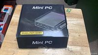 Closed retail box labeled “Mini PC Small Fast Quiet” on top of wooden surface