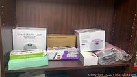 Shelf with various beauty devices and kits including a white 2-in-1 LED UV lamp box, Tomicca dip powder kit box, multifunctional polishing machine box, green heating pad box, nail dust collector box, and Wowax wax heater box.