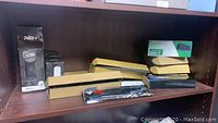Photo showing multiple cardboard boxes, a black charger box labeled PWR+, and a green box labeled X-One on a wooden shelf.
