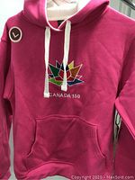 Front view of pink hooded sweater with Canada 150 multicolored maple leaf logo and white drawstrings, showing tag for size large.