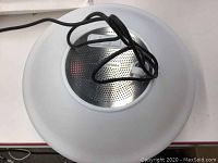 Top view of the Pool Master floating speaker, showing the silver speaker grill and white circular outer structure with USB cable attached.