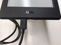 Front view of Kindle 8th generation black e-reader showing part of touchscreen and Kindle logo on frame, connected to USB charging cable.