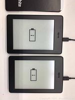 Two black Kindle Paperwhite tablets side by side with charging cables plugged in, screens white with battery icons.