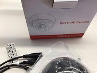 White dome surveillance camera wrapped in plastic with attached cables and connectors, next to its retail box showing camera line drawing and CCTV HD Camera branding.