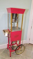 Front-left view of red and gold popcorn cart showing wheels, kettle inside, power cord